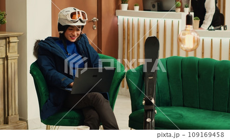 Tourist reading reviews on laptop at luxury hotel, sitting in lounge area with skiing equipment and accessories. Asian woman working remote on pc before practicing extreme winter sport in the cold. Tourist reading reviews on laptop at luxury hotel, sitting in lounge area with skiing equipment and accessories. Asian woman working remote on pc before practicing extreme winter sport in the cold. 109169458