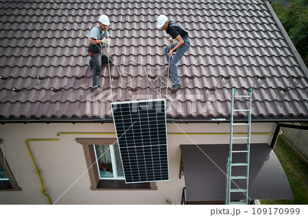 Men workers lifting up photovoltaic solar moduls on roof of house. Engineers in helmet installing solar panel system outdoors. Concept of alternative and renewable energy. Aerial view. 109170999