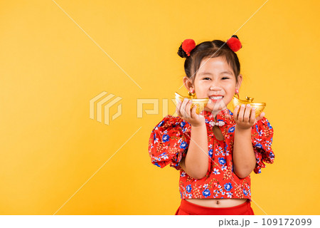 Chinese New Year. Happy Asian Chinese little girl smile wearing red cheongsam holding gold ingot, Portrait children in traditional dress hold golden bar, studio short isolated on yellow background Chinese New Year. Happy Asian Chinese little girl smile wearing red cheongsam holding gold ingot, Portrait children in traditional dress hold golden bar, studio short isolated on yellow background 109172099