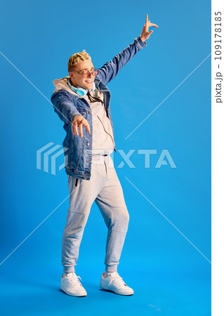 Young positive stylish guy in sunglasses, jeans jacket and casual sportswear, wit headphones, smiling against blue studio background 109178185