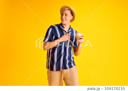 Handsome smiling young man in striped shirt, shorts and straw hat eating delicious vanilla ice cream against yellow studio background Handsome smiling young man in striped shirt, shorts and straw hat eating delicious vanilla ice cream against yellow studio background 109178210