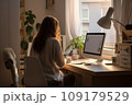 young woman working from home in front of a computer monitor. The apartment is minimalistic, with plants 109179529