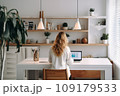 young woman working from home in front of a computer monitor. The apartment is minimalistic, with plants 109179533