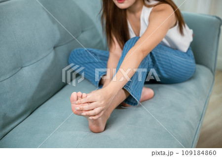 Asian beautiful female problems with foot at home, painful ankle injury, Young woman sitting on sofa holding her feet and stretch muscles have symptoms feeling pain, Health care and medical concept 109184860
