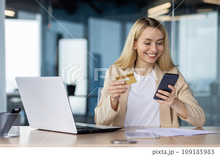 Young joyful woman at workplace inside office holds phone , bank credit debit card in hands, businesswoman chooses gifts in online store, book service and transfer funds using app on smartphone. 109185983