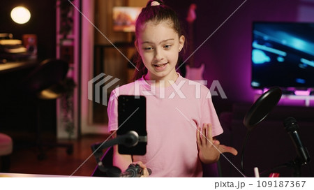 Kid films with smartphone attached to selfie stick in pink neon lit living room used as professional studio. Young media star captures footage with cellphone camera, discussing about fun day at school Kid films with smartphone attached to selfie stick in pink neon lit living room used as professional studio. Young media star captures footage with cellphone camera, discussing about fun day at school 109187367