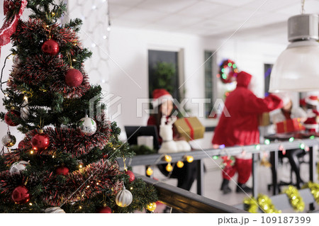 Festively decorated christmas tree close up with business workers receiving presents from Santa Claus in blurry background. Xmas adorn modern workspace during winter holiday season 109187399