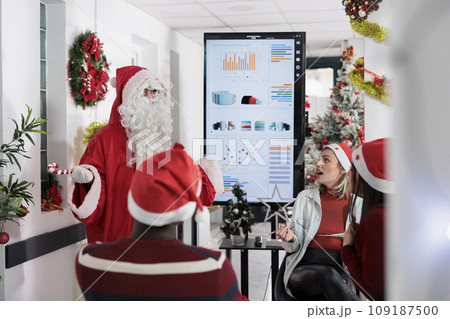 Coworker dressed as Santa Claus entertaining colleagues during business meeting in Christmas decorated workspace. Company executive having fun with coworkers in office during winter holiday season Coworker dressed as Santa Claus entertaining colleagues during business meeting in Christmas decorated workspace. Company executive having fun with coworkers in office during winter holiday season 109187500