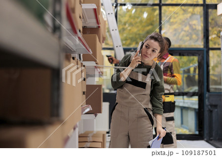 Storage room worker having remote conversation with supervisor, talking at landline phone in storage room. Woman working at products inventory, checking boxes with clients orders preparing shipment 109187501