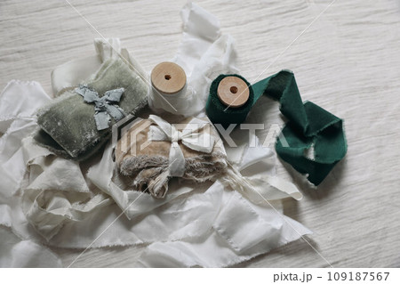 Set of silk, green and beige velvet ribbons, wooden spools on blurred muslin table cloth. Bunch of textile, sewing items. Christmas gift wrapping, craft wedding concept. Flatlay, top view, no people. Set of silk, green and beige velvet ribbons, wooden spools on blurred muslin table cloth. Bunch of textile, sewing items. Christmas gift wrapping, craft wedding concept. Flatlay, top view, no people. 109187567