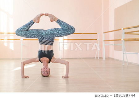Flexible Caucasian girl stands on her head in a dance class. The girl does yoga. . Woman dancing breakdance on the background of ballet barre. Flexible Caucasian girl stands on her head in a dance class. The girl does yoga. . Woman dancing breakdance on the background of ballet barre. 109189744