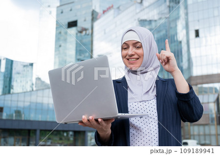Business woman in hijab and suit is using a laptop and holding her index finger up outdoors. 109189941