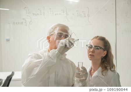 Two chemist colleagues write formulas on glass. Caucasian elderly man and young woman brainstorming. 109190044