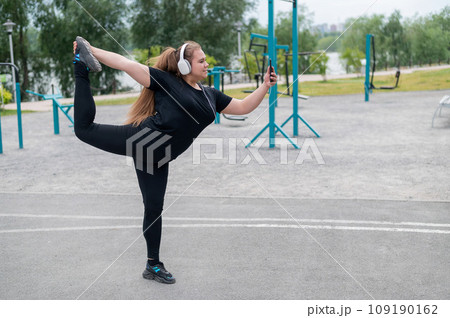 Beautiful fat young woman in headphones makes a figure skater and takes a selfie. A chubby smiling girl listens to music and warms up before jogging outdoors. Sports for weight loss. Beautiful fat young woman in headphones makes a figure skater and takes a selfie. A chubby smiling girl listens to music and warms up before jogging outdoors. Sports for weight loss. 109190162