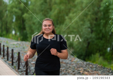 An obese young woman jogging outdoors. Fat beautiful smiling girl in a black tracksuit is engaged in fitness for weight loss on the waterfront. A woman runs on a summer day. 109190383
