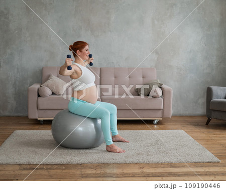 Pregnant woman doing exercises with dumbbells while sitting on a fitness ball at home. Pregnant woman doing exercises with dumbbells while sitting on a fitness ball at home. 109190446