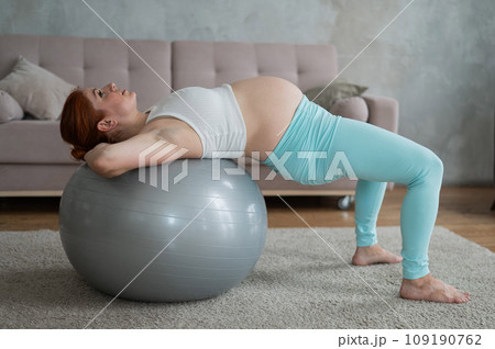 Pregnant red-haired woman doing exercises on fitness ball at home.  109190762