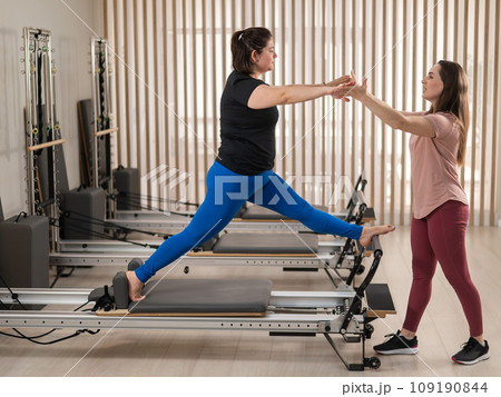 Overweight caucasian woman doing pilates exercises on reformer with personal trainer.  109190844