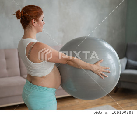 Pregnant red-haired woman doing arm exercises with fitball at home. 109190883