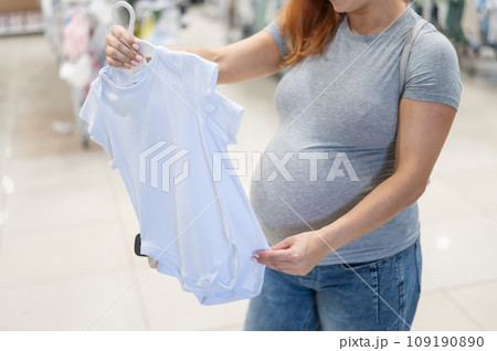 Caucasian pregnant woman chooses baby clothes in a store. Faceless expectant mother in the 3rd trimester.  109190890