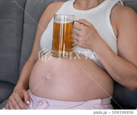 A faceless pregnant woman sits on the sofa and holds a glass of beer on her stomach. 109190956