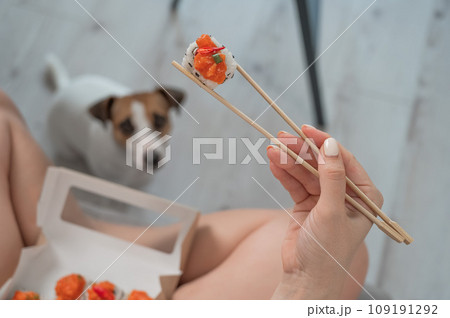 A woman sits on the sofa and eats rolls. Jack Russell Terrier dog sits on the floor and begs for food from its owner.  109191292