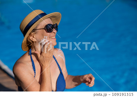 Portrait of an old woman in a straw hat, sunglasses and a swimsuit applying sunscreen to her face while relaxing by the pool.  109191315