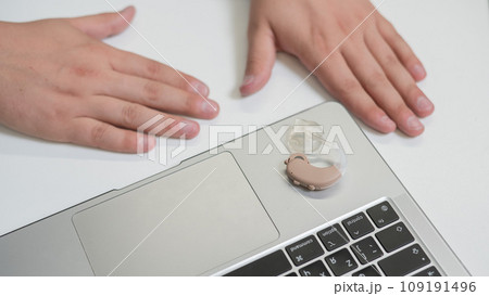 Close-up of a man's hands with a hearing aid near a laptop.  109191496
