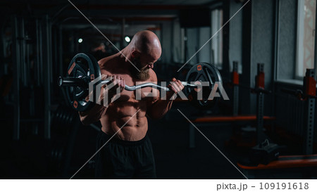 Caucasian bald topless man doing an exercise with a barbell in the gym. Bicep curls with weights. Caucasian bald topless man doing an exercise with a barbell in the gym. Bicep curls with weights. 109191618