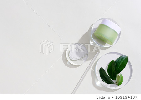 A jar of eye cream is placed on a glass platform, a few green tea leaves are decorated next to it on a white background. Green tea essence helps reduce eye puffiness and eliminate dark circles. A jar of eye cream is placed on a glass platform, a few green tea leaves are decorated next to it on a white background. Green tea essence helps reduce eye puffiness and eliminate dark circles. 109194187