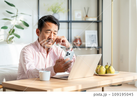 リビングでノートパソコンとスマホを使うシニア男性。 リビングでノートパソコンとスマホを使うシニア男性。 109195045