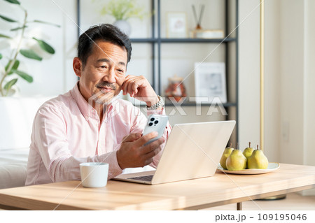 リビングでノートパソコンとスマホを使うシニア男性。 109195046