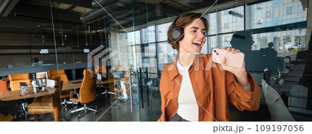 Image of happy young digital nomad, woman in office, dancing and singing, listening music on smartphone with wireless headphones Image of happy young digital nomad, woman in office, dancing and singing, listening music on smartphone with wireless headphones 109197056