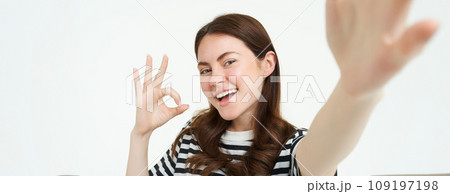 Portrait of positive, happy young woman shows okay sign and takes selfie, holds smartphone with extended hand, posing near something she recommends, isolated on white background 109197198