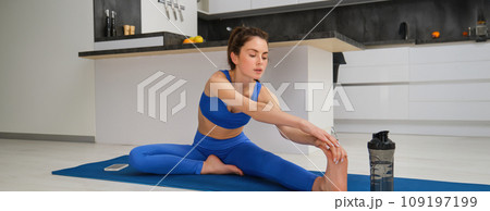 Fitness and workout concept. Young woman stretching her legs, doing exercises at home on yoga mat, doing splits on floor in living room Fitness and workout concept. Young woman stretching her legs, doing exercises at home on yoga mat, doing splits on floor in living room 109197199