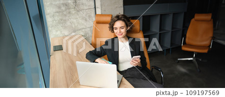 Portrait of young professional, businesswoman sitting in her office, answering phone call, using her smartphone and laptop, working near window Portrait of young professional, businesswoman sitting in her office, answering phone call, using her smartphone and laptop, working near window 109197569