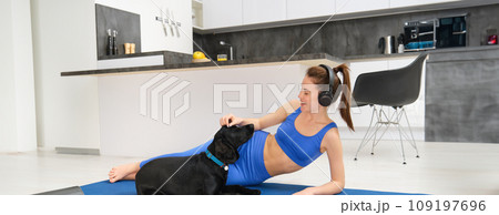 Portrait of happy young woman, tried to workout at home, laying on yoga mat in activewear, playing with her dog, listening music in wireless headphones 109197696