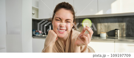 Portrait of beautiful brunette girl cooking in the kitchen, posing in bathrobe at home, holding zucchini, showing happy smile, making healthy food, vegetarian meal 109197709