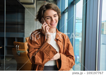 Happy young office worker near window, calling someone, answers a call on mobile phone, talking and smiling Happy young office worker near window, calling someone, answers a call on mobile phone, talking and smiling 109197767