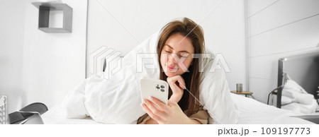 Portrait of beautiful brunette woman with smartphone, relaxing in her bed under blankets and smiling, checking social media feed on mobile phone app, wrap herself in duvet and enjoying lazy morning 109197773