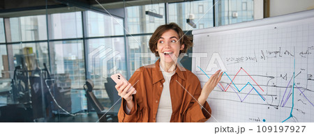 Portrait of woman expresses excitement, office worker dances near diagram with company stats, holds smartphone, reacts to wonderful news or achievement 109197927