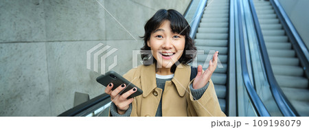 Happy triumphing asian girl, looks amazed and joyful, holds smartphone, goes down escalator with pleased face, wins, hears good news Happy triumphing asian girl, looks amazed and joyful, holds smartphone, goes down escalator with pleased face, wins, hears good news 109198079