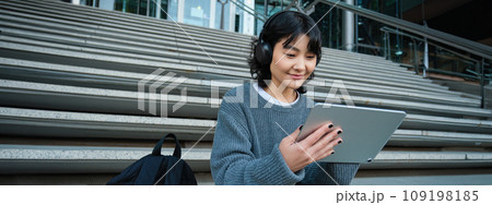 Young asian girl, student in headphones, works on remote, digital artist drawing on tablet with graphic pen, listening music in headphones and sitting on street staircase 109198185