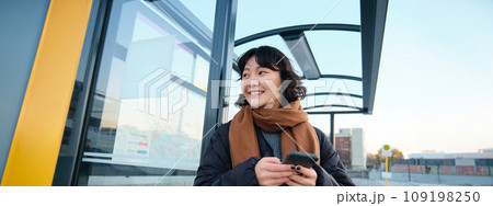 Cute smiling asian girl standing on bus stop, holding smartphone, wearing winter jacket and scarf. Woman commuting to work or university via public transport, stands on road Cute smiling asian girl standing on bus stop, holding smartphone, wearing winter jacket and scarf. Woman commuting to work or university via public transport, stands on road 109198250