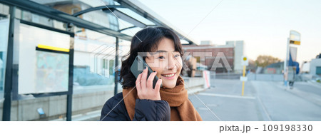 Smilling Korean girl talking on mobile phone, standing on bus stop, using smartphone, posing on road in winter, wrapped in scarf, wearing black jacket 109198330