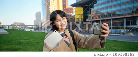 Happy university student, korean girl takes selfie with her papers and digital tablet, holds smartphone and poses near university campus 109198365