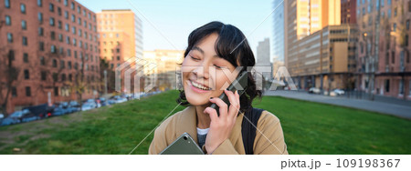 University lifestyle. Young asian student, girl with tablet and notebook, talks on mobile phone while walks from campus on street 109198367