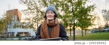 Smiling young girl, korean student sits in park in scarf and hat on chilly day, draws outdoors on digital tablet with graphic pen, makes scatches Smiling young girl, korean student sits in park in scarf and hat on chilly day, draws outdoors on digital tablet with graphic pen, makes scatches 109198442