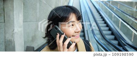 Headshot of smiling korean woman with smartphone, makes a phone call, goes down escalator in city, commutes to university Headshot of smiling korean woman with smartphone, makes a phone call, goes down escalator in city, commutes to university 109198590