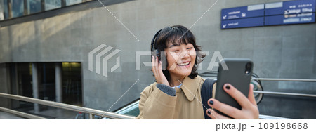 Beautiful korean girl in headphones, takes selfie on her smartphone, stands on street with mobile phone, records video, listens music and travels around city Beautiful korean girl in headphones, takes selfie on her smartphone, stands on street with mobile phone, records video, listens music and travels around city 109198668